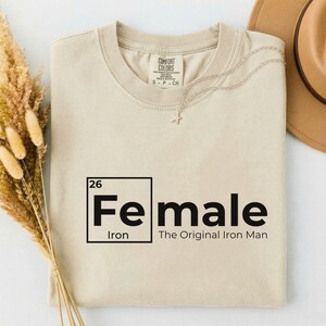 May include: Beige Comfort Colors t-shirt with the text "Female" in a black, periodic table-style design, with "Iron" and "The Original Iron Man" below. A silver necklace with a star pendant rests on the shirt.