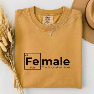 May include: Mustard-colored t-shirt with the text "Fe male" in a black, bold font, with the periodic symbol for iron. The phrase "The Original Iron Man" is below the text. A silver star pendant necklace and a tan hat are also visible.