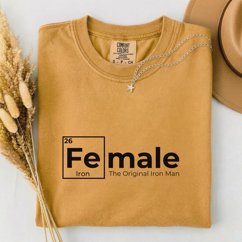 May include: Mustard-colored t-shirt with the text "Fe male" in a black, bold font, with the periodic symbol for iron. The phrase "The Original Iron Man" is below the text. A silver star pendant necklace and a tan hat are also visible.