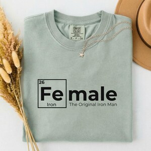 May include: A light green t-shirt with the text "Fe male" in a black, periodic table-style box, with "Iron" below. Below the box is the text "The Original Iron Man". A gold necklace with a star pendant is on the shirt.