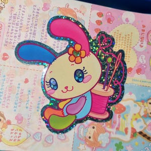 Könnte beinhalten: Ein glitzernder Aufkleber mit einem Cartoon-Hasen mit großen blauen Augen, gelbem Kopf und blauem Körper. Der Hase hat ein blaues und ein rosa Ohr, eine kleine orangefarbene Blume auf dem Kopf und sitzt neben einer rosa Garnrolle mit zwei Nadeln.