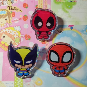 Puede incluir: Un conjunto de pegatinas de personajes de dibujos animados, incluyendo a Deadpool, Wolverine y Spider-Man. Las pegatinas son coloridas con un efecto de purpurina holográfica. El fondo incluye texto e ilustraciones de fresas y personajes de dibujos animados.