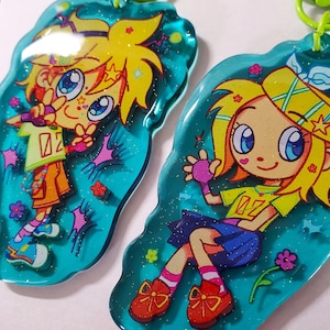 May include: Two cartoon character keychains. Each keychain features a colorful anime-style character with yellow hair, wearing a green shirt with the number "02". The keychains are outlined in teal and have a green clasp.