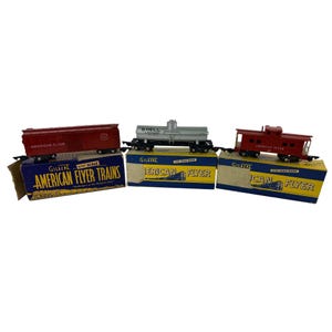 May include: Three vintage American Flyer train cars, including a red boxcar, a silver Shell tanker car, and a red caboose, displayed on their original boxes. The boxes are blue and yellow with the words "American Flyer Trains" printed on them.