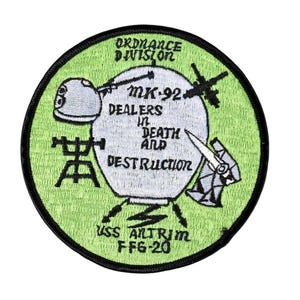 May include: A circular patch with a green border and background. The patch features the text "ORDNANCE DIVISION" at the top and "MK-92 DEALERS IN DEATH AND DESTRUCTION" in the center. The bottom of the patch reads "USS ALTRIM FFG-20".