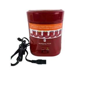 May include: A burgundy Wolfgang Puck electric cooker with a black power cord. The cooker has a label with text about preparing meals, including rice, chicken soup, and chocolate cake. It has cook and warm indicator lights.