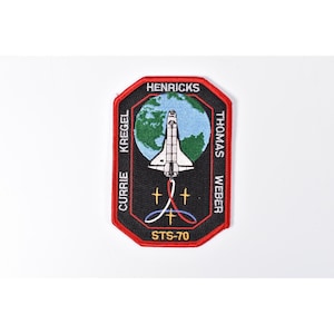 May include: An embroidered mission patch with a black background and red border. It features a white space shuttle flying in front of a blue and green Earth. The text reads HENRICKS, KREGEL, CURRIE, THOMAS, WEBER, and STS-70, with gold stars.