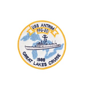 May include: A circular patch with a yellow border and white background. The patch features a ship illustration, the text "USS ANTRIM FFG-20", "GREAT LAKES CRUISE", and "1988". The ship is depicted in white and gray, with blue water and land details.