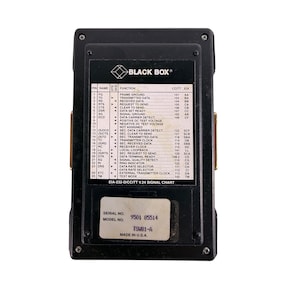 May include: Black Box EIA-232-D/CCITT V.24 signal chart. The black rectangular device has a label with the Black Box logo and technical specifications. The serial number is 9501 85514, and the model number is TSMB1-A. Made in the U.S.A.