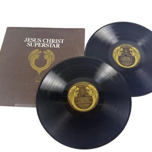May include: A vintage vinyl record album titled "JESUS CHRIST SUPERSTAR" with two black records and a brown album cover. The record labels are gold with black text and a decorative design. The album cover has the title and a gold emblem.