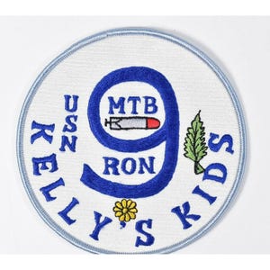 May include: Embroidered patch with a white background and a light blue border. The patch features the text "KELLY'S KIDS" in blue, along with "USN" and "MTB RON 9". A stylized torpedo and a yellow flower are also present.