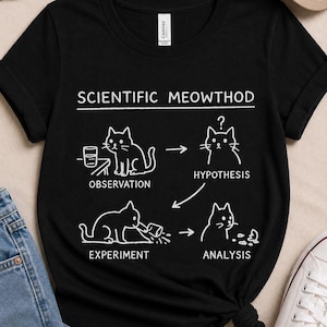Puede incluir: Camiseta negra con un gráfico blanco que ilustra el método científico utilizando gatos. El texto dice "Scientific Meowthod" encima del diagrama, que incluye pasos como "Observación" e "Hipótesis".