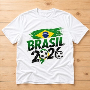 Puede incluir: Camiseta blanca con la bandera brasileña y la inscripción "BRASIL 2026" en verde y negro. El diseño incluye balones de fútbol, sugiriendo un tema deportivo.