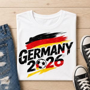 Op de afbeelding: Wit T-shirt met de tekst "GERMANY 2026" in zwart, met een voetbalafbeelding. Het ontwerp bevat de kleuren van de Duitse vlag: zwart, rood en geel. Het shirt wordt getoond met een blauwe jeans en zwarte sneakers.