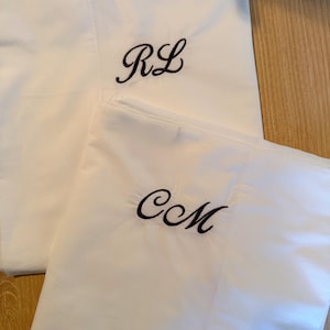 May include: Two white pillowcases with embroidered initials. One pillowcase features the initials "RL" in black script, while the other displays "CM". The pillowcases are folded and placed on a light-coloured wooden surface.