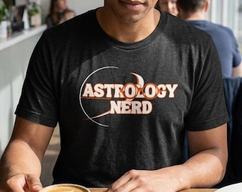 May include: A black t-shirt with the words "ASTROLOGY NERD" in orange and white lettering. The person is holding a ceramic mug with a latte. A notebook and pen are on the wooden table.