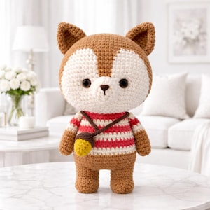 May include: A handmade crochet fox doll with brown, white, and red striped details. The fox has large black eyes, brown ears, and a small yellow acorn bag. The doll is standing on a marble table in a well-lit room.