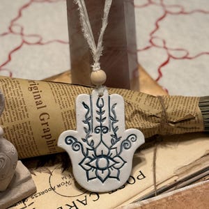 May include: White ceramic Hamsa hand pendant with a dark blue floral design, hanging from a white string with a wooden bead. A clear, pink crystal and vintage paper are in the background.