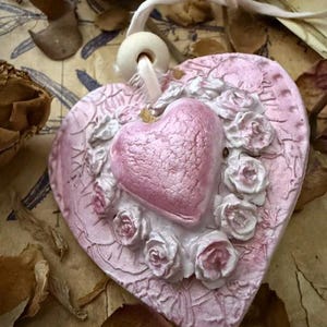 May include: A pink heart-shaped ornament with a smaller heart in the center, surrounded by white and pink roses. The ornament has a textured surface and a white string for hanging. The background includes dried leaves and paper.
