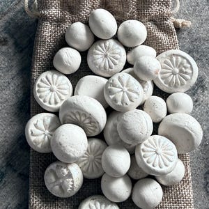 May include: A collection of white, decorative stones in various shapes, including spheres and flat, circular pieces with floral designs. The stones are inside a small, beige burlap drawstring bag, with the bag's texture visible. The background is a textured gray surface.