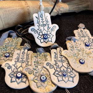 May include: A collection of white ceramic Hamsa hand ornaments with blue and gold accents. Each hand features an eye symbol and floral designs. Some have blue ribbons for hanging. The ornaments are arranged on a dark surface.