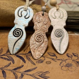 May include: Three pendant necklaces with a goddess figure design. The pendants are in silver, bronze, and white, each with a spiral detail. They hang on cords with wooden beads, set against a vintage paper background.