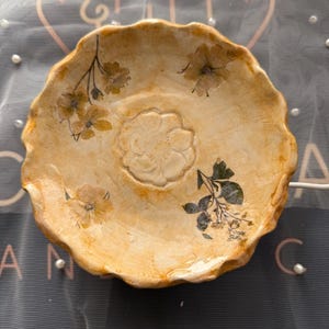 May include: A round ceramic dish with a scalloped edge and a glossy, mottled yellow and cream finish. The center features a raised floral relief, while the surface is decorated with pressed botanical illustrations of yellow and dark green flowers.