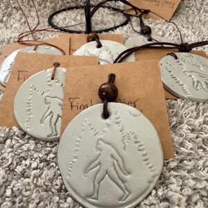May include: Pale green ceramic pendants with a Bigfoot design, strung on brown cords. Each pendant is round with a raised image of Bigfoot walking through a forest. The pendants are attached to brown paper tags.
