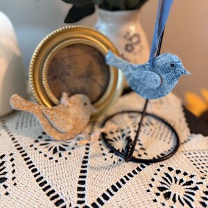 May include: Two decorative bird ornaments, one gold and one blue, rest on a cream-colored lace doily. A small gold-framed picture and a black metal stand are also visible. The background includes a vase with greenery.