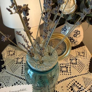 May include: A blue glass jar topped with a textured, light-colored clay flower frog featuring multiple holes. Several dried lavender stems are inserted into the clay frog. A white card with printed text is visible in the lower left corner.