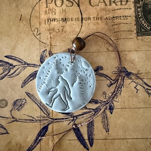 May include: A light blue pendant featuring a Bigfoot design walking through a forest, with a moon in the background. The pendant is round and has a brown wooden bead attached to a brown cord. The background is a vintage postcard.
