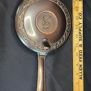 May include: A vintage bronze-toned hand mirror with an ornate, floral-like border. The mirror's face features a circular design with the letter "S" in the center. The handle is a continuation of the mirror's frame. A wooden ruler is next to the mirror.