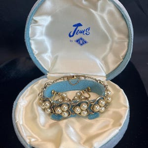 May include: Vintage jewelry set in a light blue velvet box. The set includes a gold-toned bracelet and matching clip-on earrings, each adorned with faux pearls. The box is lined with satin and features the text "Jems by MS".