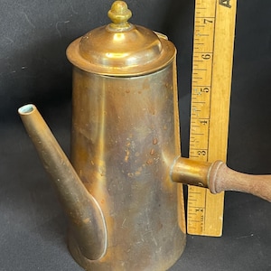 May include: A brass coffee pot with a wooden handle and a lid, positioned next to a yellow wooden ruler showing a height of approximately 8 inches. The metal surface displays a weathered patina with visible spots and a cylindrical body shape.