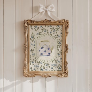 May include: A framed watercolour print featuring a teapot with blue floral details. The print is set against a backdrop of blue flowers and green leaves. The ornate gold frame is suspended by a white ribbon and bow.