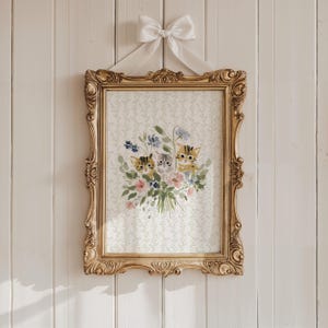 Puede incluir: Una impresión en acuarela enmarcada que presenta tres gatitos entre un ramo de flores. La obra de arte está enmarcada en un marco dorado ornamentado y está suspendida por una cinta blanca atada con un lazo. El fondo es un patrón floral claro.