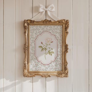 May include: A framed floral print with a gold ornate frame. The print features a light pink rose illustration on a patterned background of small pink flowers. A white ribbon is tied at the top for hanging.