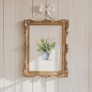 Puede incluir: Una impresión en acuarela enmarcada de flores de lirios de los valles en un jarrón azul con lunares blancos. La impresión está en un marco dorado adornado y está suspendida por una cinta blanca atada en un lazo. El fondo es una pared de madera blanca.