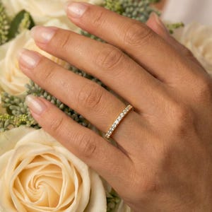 Może przedstawiać: Złoty pierścionek z małymi, przezroczystymi kamieniami. Pierścionek jest na palcu, w tle bukiet białych róż. Klasyczny design, często noszony jako obrączka ślubna lub pierścionek zaręczynowy.