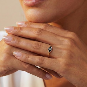 Può includere: Un anello d'oro con un design a forma di diamante in smalto nero e una piccola gemma bianca. L'anello è indossato su un dito, evidenziando i suoi dettagli delicati e il suo stile elegante. Il design dell'anello è semplice ma accattivante.
