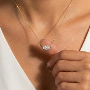 Puede incluir: Un delicado collar de oro con un colgante en forma de marquesa con tres piedras preciosas transparentes. El colgante está centrado en una fina cadena de oro. El collar se lleva sobre una parte superior blanca.