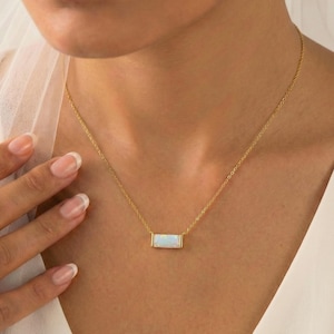 Op de afbeelding: Een goudkleurige ketting met een rechthoekige, lichtgekleurde hanger, mogelijk een opaal. De fijne ketting rust op de borst. De achtergrond is een zachte, neutrale kleur. De hanger is ongeveer 1,5 cm lang.