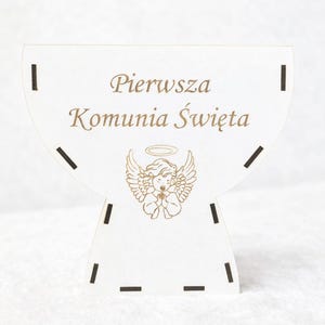 Puede incluir: Decoración de madera blanca cortada con láser con el texto polaco "Pierwsza Komunia Święta" y una ilustración de ángel. La decoración tiene una forma única con detalles recortados y está diseñada para colocarse de pie.