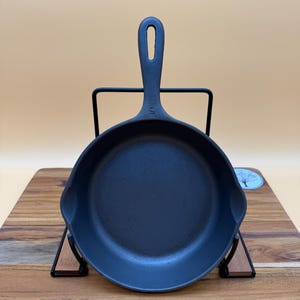 May include: A black cast iron skillet with a long handle and a loop for hanging. The skillet is displayed on a black metal stand and a wooden cutting board. The number "5" is visible on the handle. A tag with the text "5WU1225" is attached.