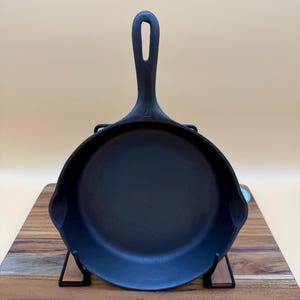 May include: A black cast iron skillet with a long handle and a rounded cooking surface. The skillet is displayed on a wooden cutting board and supported by a black metal stand. The handle has a hole for hanging. A tag with the number "7WU1209" is attached.
