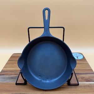 May include: A black cast iron skillet with a handle and two side spouts. The skillet is displayed on a black metal stand and a wooden cutting board. A tag with the text "5GS 1216" is attached.