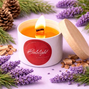 Puede incluir: Una vela encendida en una lata blanca con tapa de madera. La vela tiene cera naranja y una etiqueta roja que dice "Belolight". Está rodeada de lavanda, agujas de pino y piñas sobre una superficie púrpura.