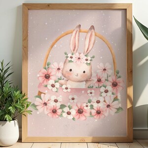 Könnte beinhalten: Ein gerahmter Wandkunstdruck mit einer Aquarellillustration eines hellbraunen Hasen, der in einem geflochtenen Korb sitzt. Der Hase trägt einen Blumenkranz und der Korb ist mit rosa und weißen Blumen vor einem zartrosa Hintergrund gefüllt.