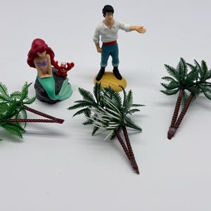May include: A set of plastic figurines including a mermaid with red hair and a green tail, a small red crab, a male figure in a white shirt and blue pants, and three miniature palm trees with brown trunks and green fronds on a white surface.