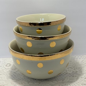 May include: A set of three nested off-white ceramic bowls featuring a metallic gold polka dot pattern and a wide gold rim. The bowls are stacked vertically, with the smallest bowl on top and the largest bowl on the bottom, showcasing a consistent design.